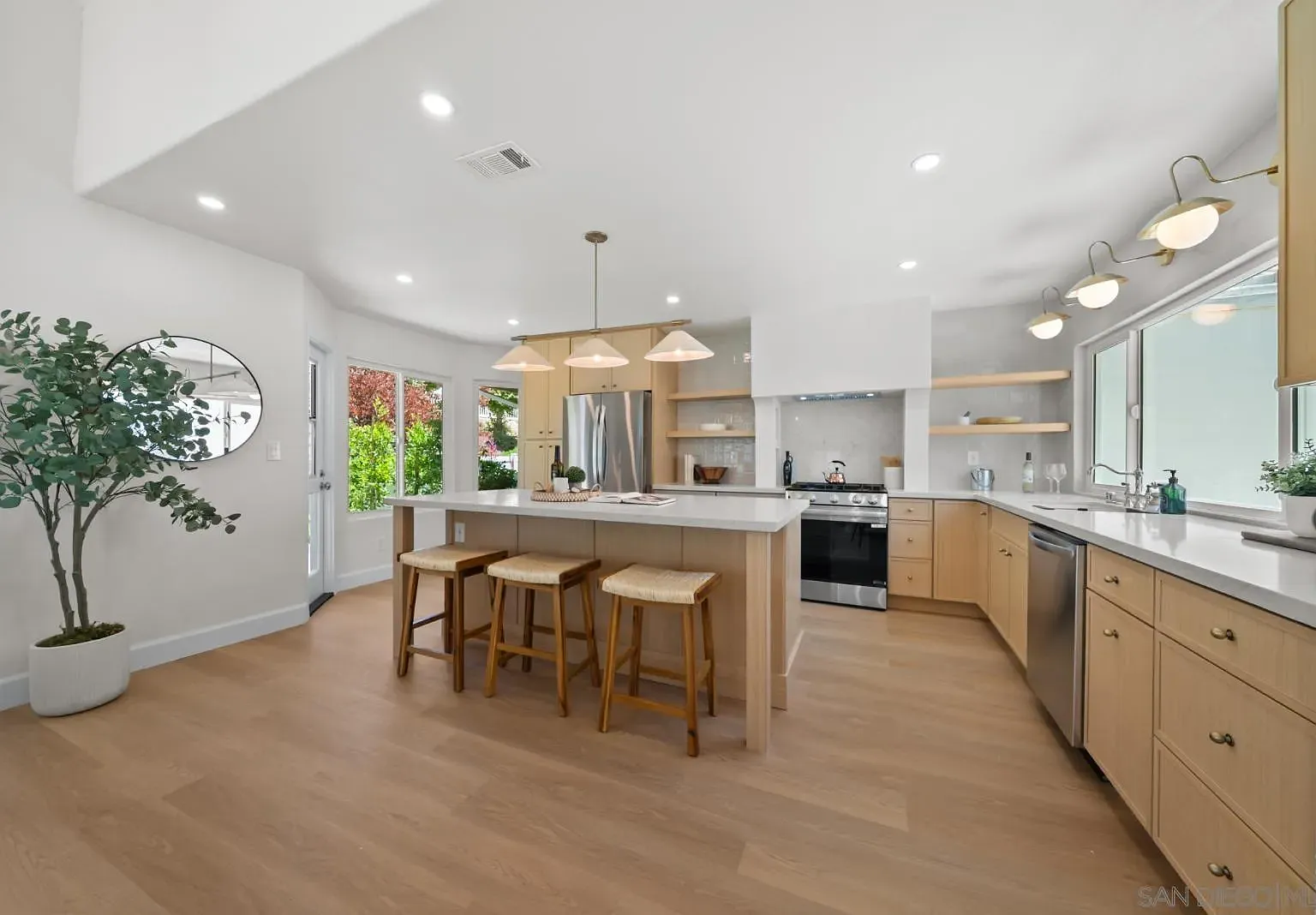 The remodeled kitchen at 4742 Collinos Way, showing the bleached oak island with three woven stools, brass pendant lights, and a faux olive tree at the entryway