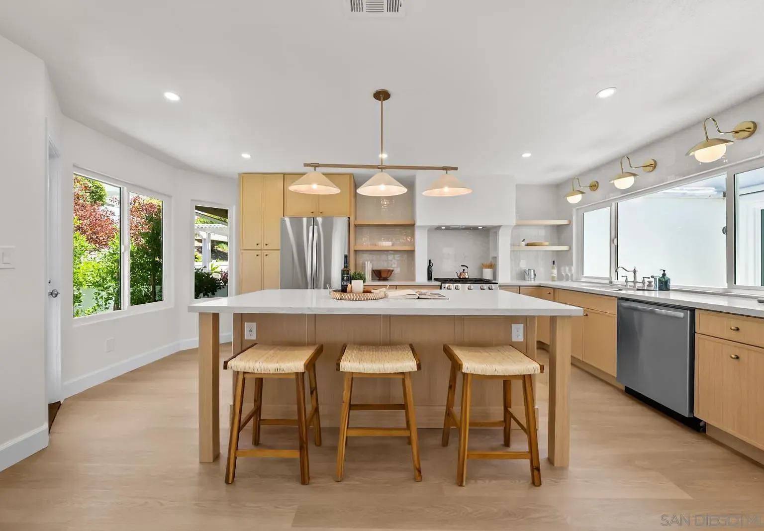 Head-on view of the kitchen island at 4742 Collinos Way with three pendant lights, woven counter stools, and brass wall sconces beside the sink